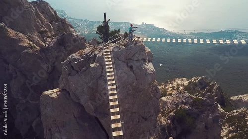 Apic aerial shot of man walking suspension bridge to Cross in Crimea. High rocks Ai-Petri of Crimean mountains. Black sea coast city and blue sky with clouds.