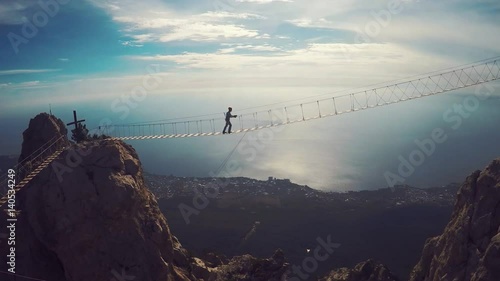 Apic aerial shot of man walking suspension bridge to Cross in Crimea. High rocks Ai-Petri of Crimean mountains. Black sea coast city and blue sky with clouds. photo