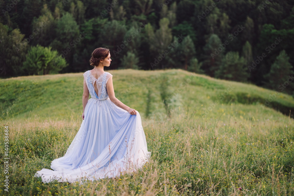 Beautiful young caucasian stylish girl in blue evening gown standing on the background of spectacular nature, hills and trees. Woman portrait outdoors. Copy space. Beauty, health care concept.