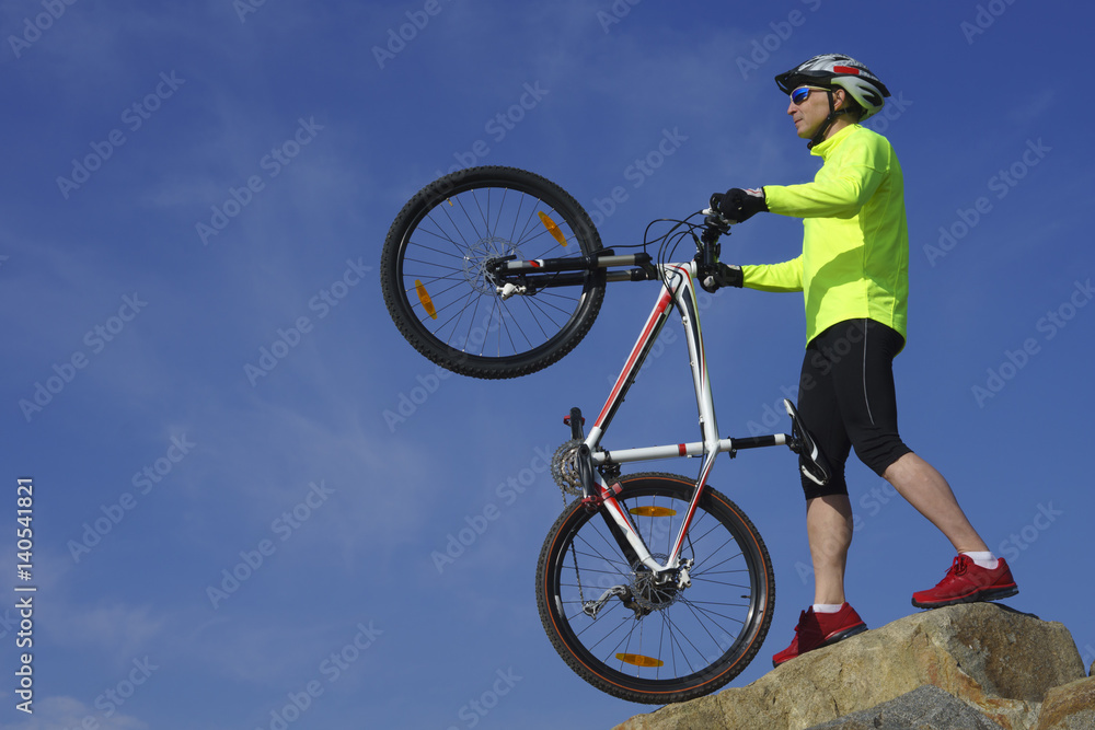 Fototapeta premium Man, in light-green jacket and red sneakers, near mountain bike with sky background