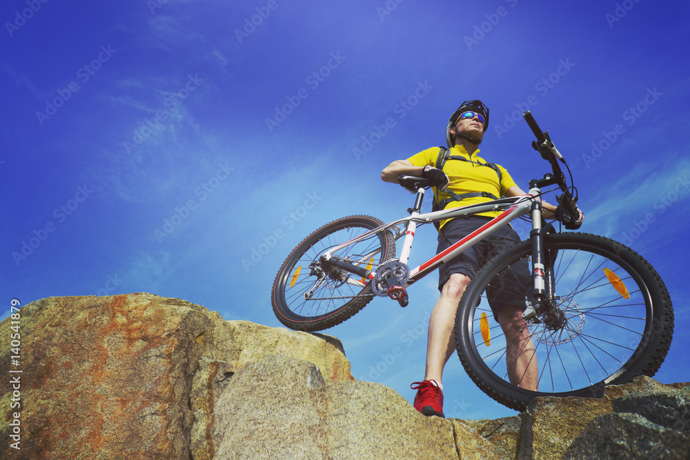 Man, in yellow t-shirt and red sneakers, near mountain bike with sky background.