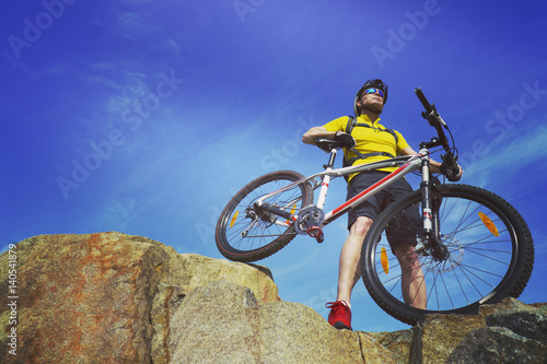 Man, in yellow t-shirt and red sneakers, near mountain bike with sky background.