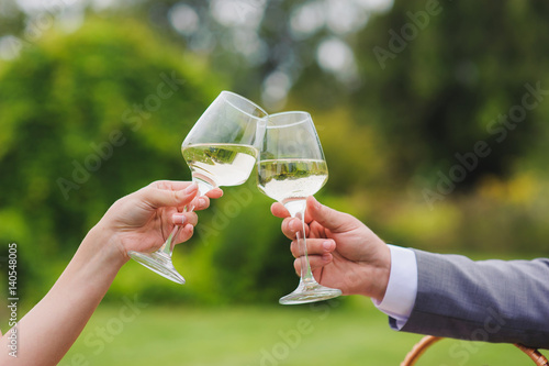 Couple drinking white wine outdoors. Clinking glasses. Grass, trees and bushes in the background.