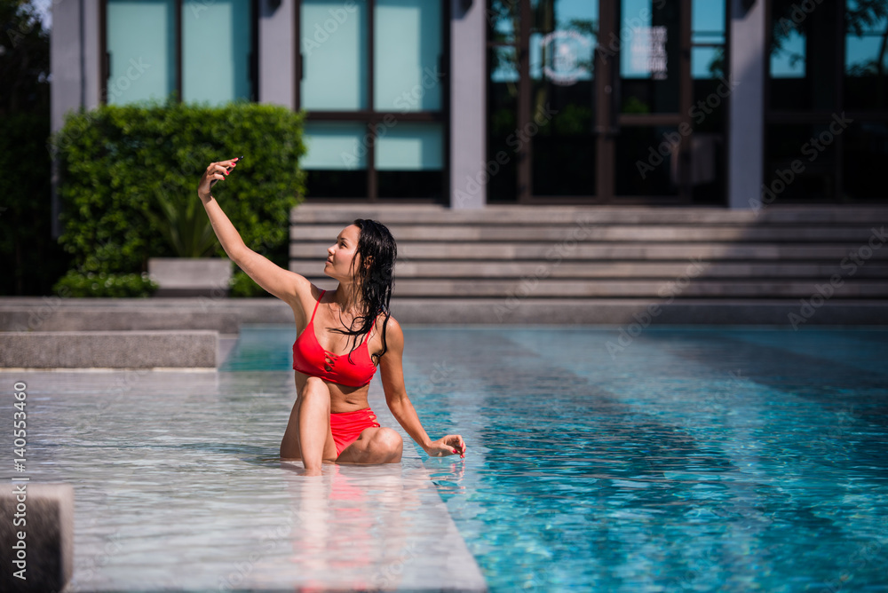 Naklejka premium Beautiful woman in bikini taking a selfie by pool side on a sunny day at luxury hotel