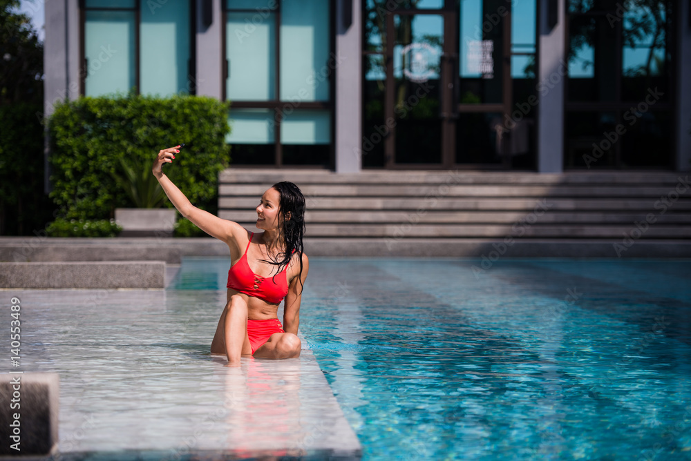 people, technology, travel, tourism and summer concept - happy young woman in bikini swimsuit and sunglasses taking selfie with smatphone over swimming pool on beach with palm trees background people, technology, travel, tourism and summer concept - happy young woman in bikini swimsuit and sunglasses taking selfie with smatphone over swimming pool on beach with palm trees background