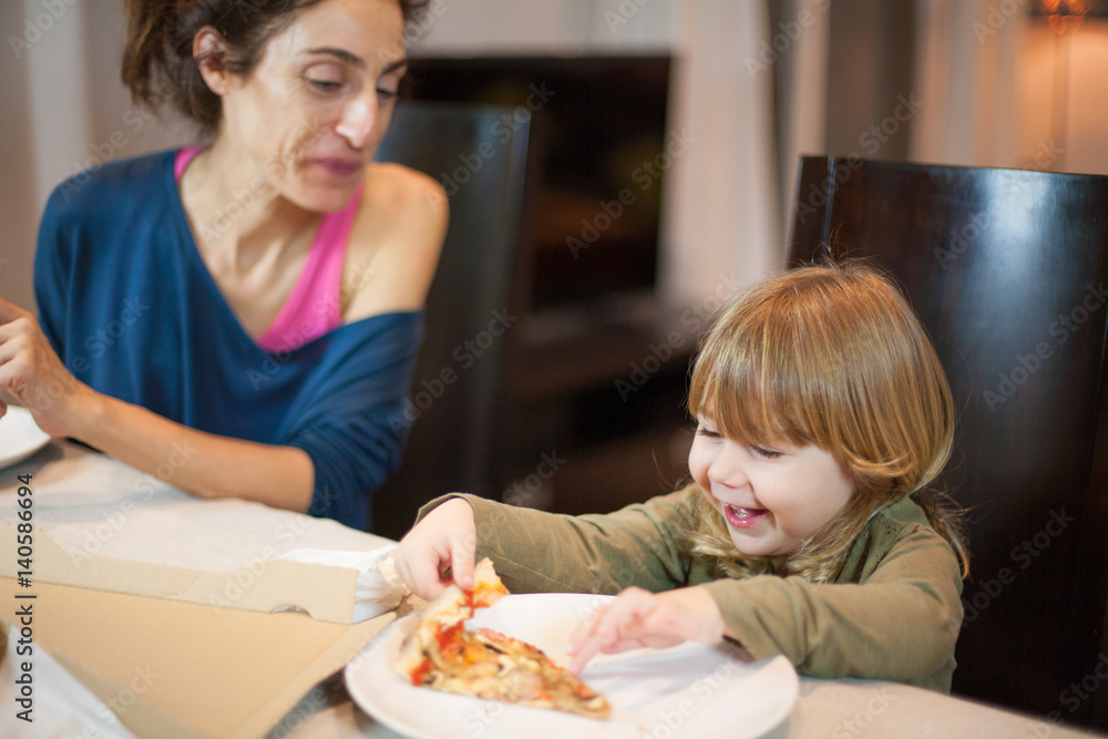 Three years old blonde child laughing taking a pizza piece, sitting in dark brown chair, besides her mother happy in table with beige tablecloth at home
