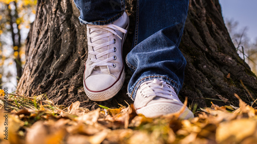 A man in white sneakers stands leaning against a big tree in an autumn park.