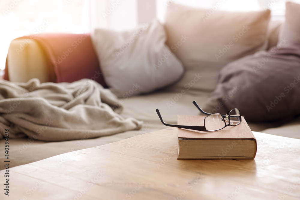 book with eyeglasses on table in front of couch - obrazy, fototapety, plakaty 