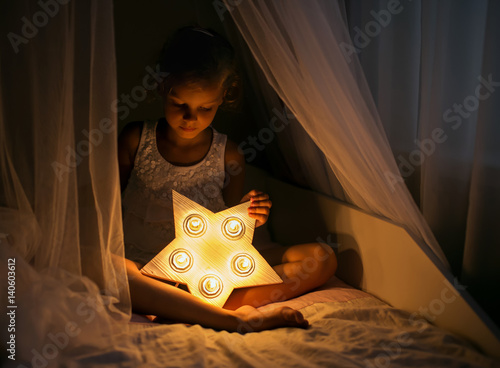 Obraz na plátně A girl is holding a  nightlight in the form of a star and is sitting on the bed