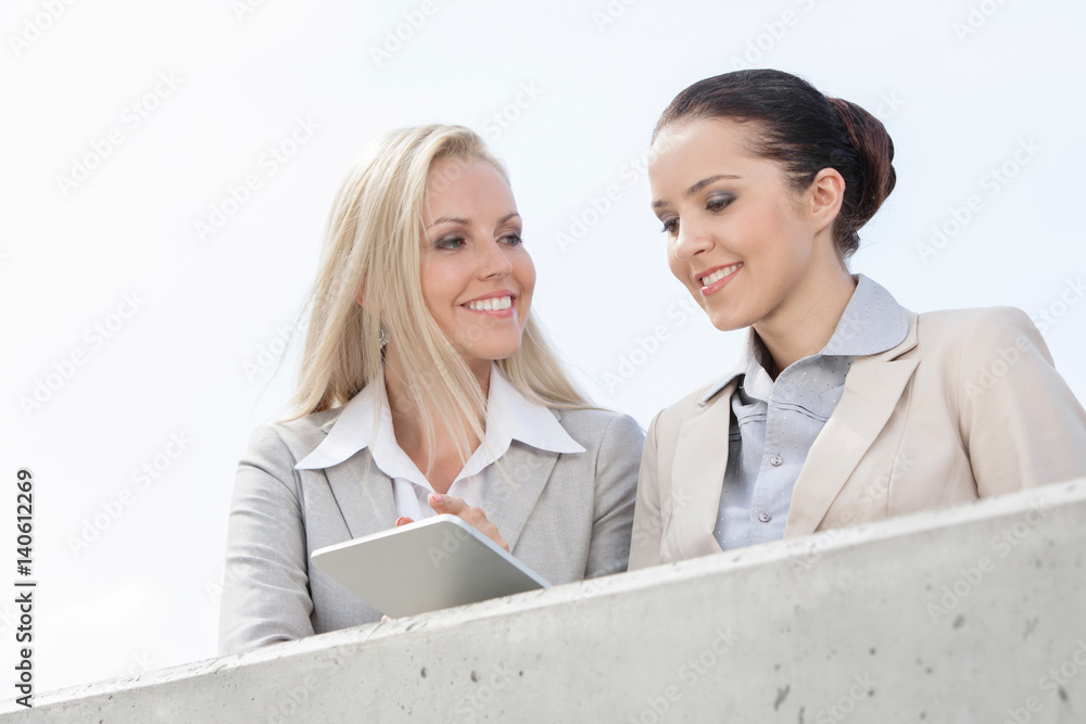 Low angle view of smiling businesswomen using digital tablet while standing on terrace against sky