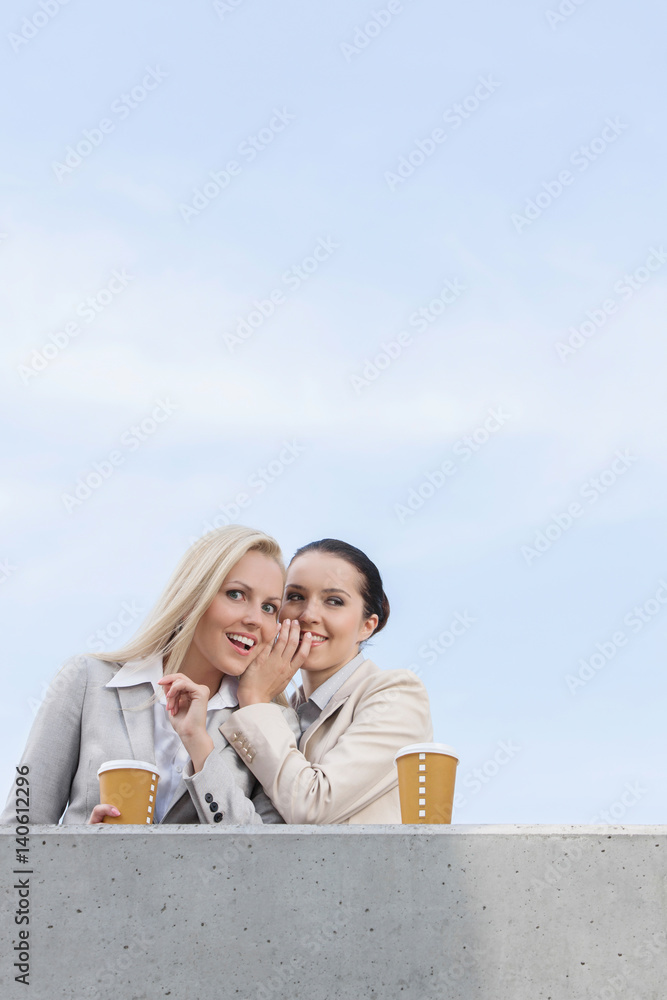 Low angle view of happy businesswoman whispering in coworker's ear while standing on terrace against sky
