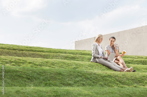 Full length of businesswomen with disposable coffee cup and laptop sitting on grass steps against sky