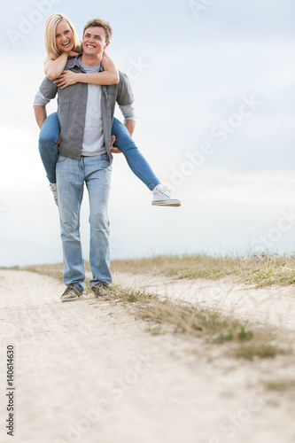 Full length of cheerful young man piggybacking woman on trail at field