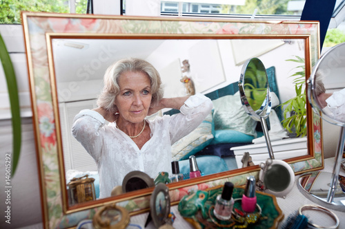 Mirror reflection of senior woman putting on necklace at home photo