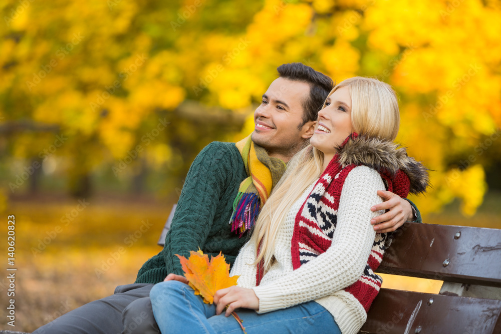Fototapeta premium Happy young couple looking away while sitting on park bench during autumn
