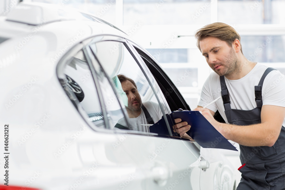 Automobile mechanic writing on clipboard while examining car in repair shop