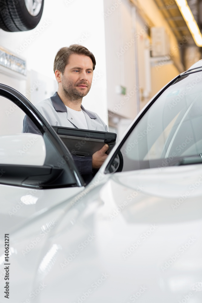 Maintenance engineer holding tablet PC while examining car in repair shop