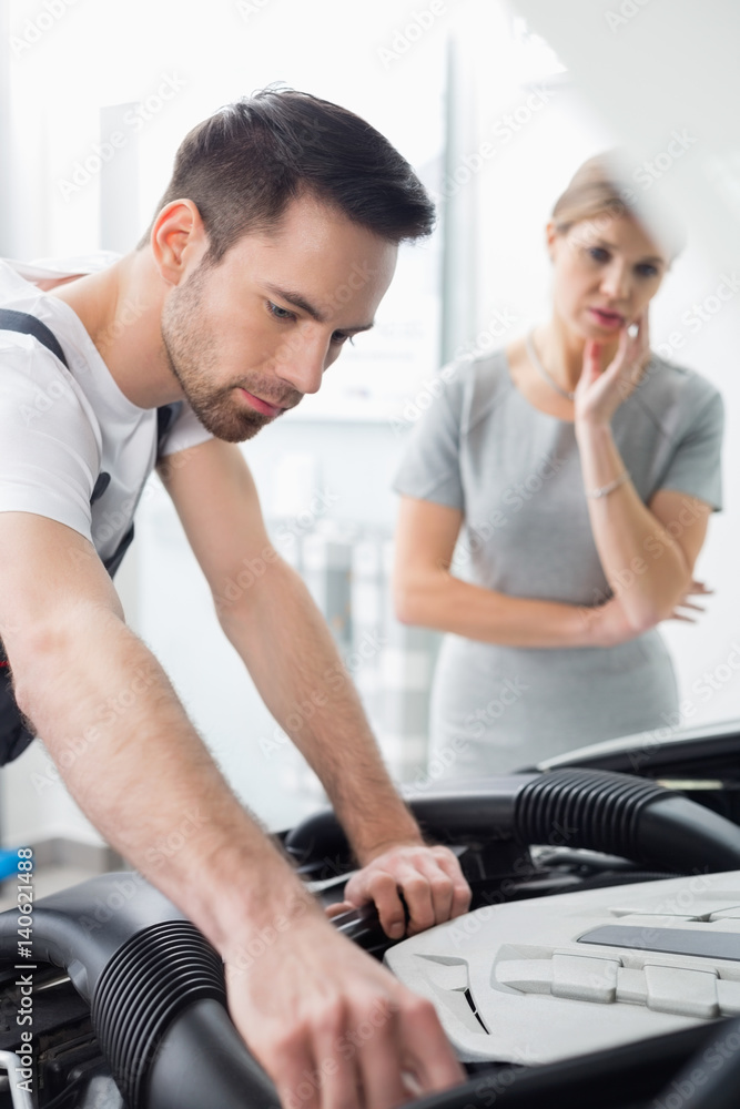 Young male technician examining car engine with worried customer in background at workshop Young male technician examining car engine with worried customer in background at workshop