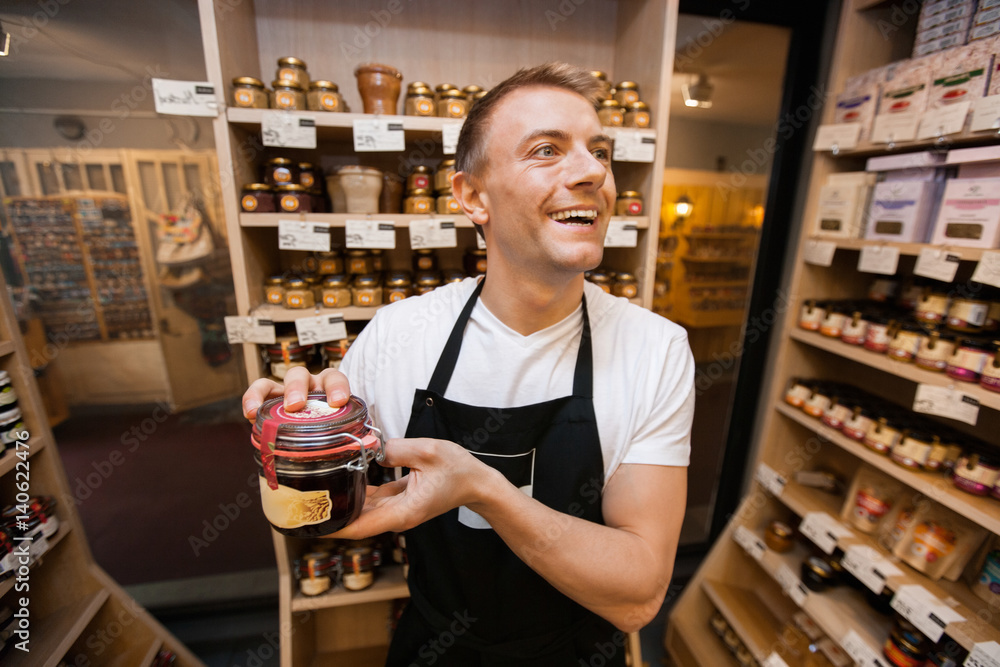 Cheerful salesman holding jar of jam in grocery store