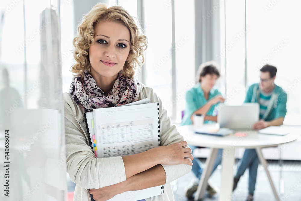Portrait of creative businesswoman holding files with colleagues working in background at office Portrait of creative businesswoman holding files with colleagues working in background at office