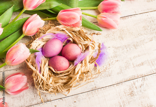 Easter eggs in nest, fresh pink tulips on white wooden background.