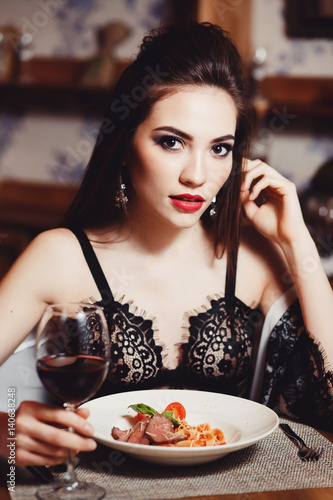 A beautiful girl in her underwear eats spaghetti pasta with meat and wine in a cafe-restaurant. She eats her hands. Dark background. Fashion. Portrait. Close-up.