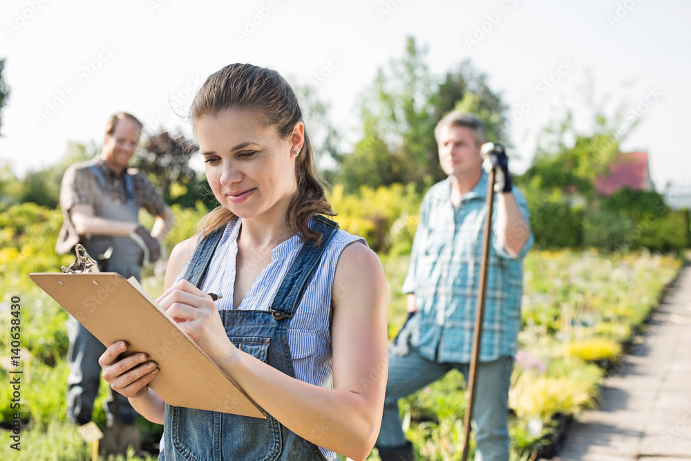 Beautiful supervisor writing on clipboard with gardeners standing in background at plant nursery Beautiful supervisor writing on clipboard with gardeners standing in background at plant nursery