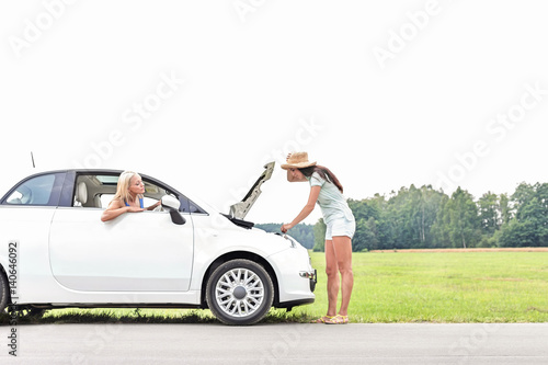 Woman looking at friend repairing broken down car on country road