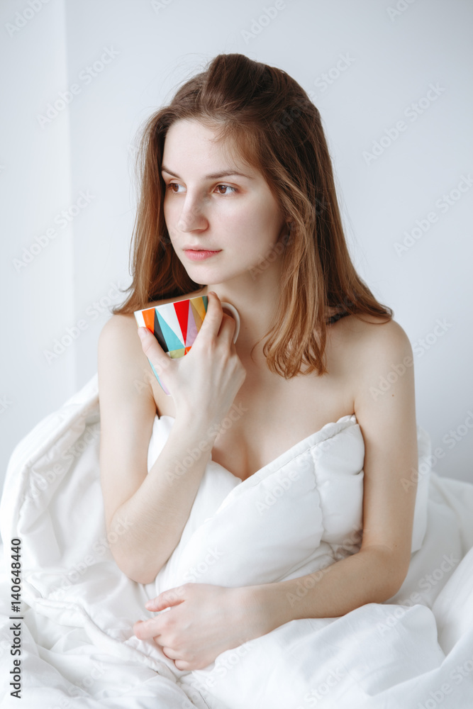 Portrait of pensive sad beautiful young Caucasian girl woman with long red hair sitting in bed wrapped covered with blanket, holiding cup, drinking coffee or tea, on white background. Portrait of pensive sad beautiful young Caucasian girl woman with long red hair sitting in bed wrapped covered with blanket, holiding cup, drinking coffee or tea, on white background.