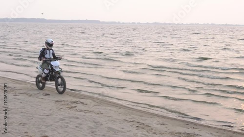 The motorcyclist riding a sport bike on a sand beside the lake at sunset. photo