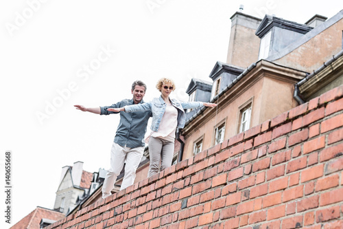 Low angle view of middle-aged couple with arms outstretched walking on brick wall against clear sky