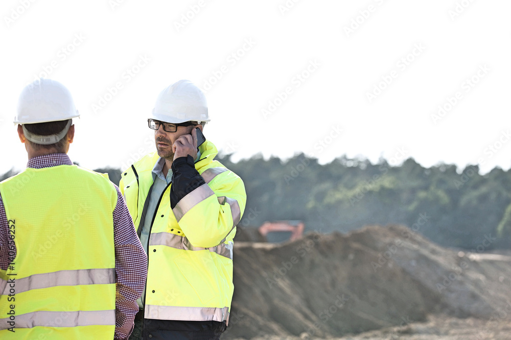 Supervisor using mobile phone while standing with colleague at construction site against clear sky Supervisor using mobile phone while standing with colleague at construction site against clear sky