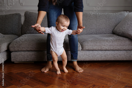 Smiling baby learning to walk in the living room with his mother holding his hands