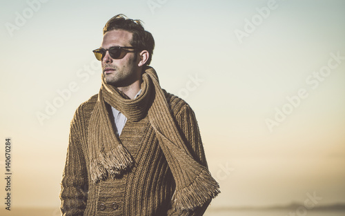 Attractive man wearing sunglasses, scarf and a brown sweater. Male model posing in mountain.