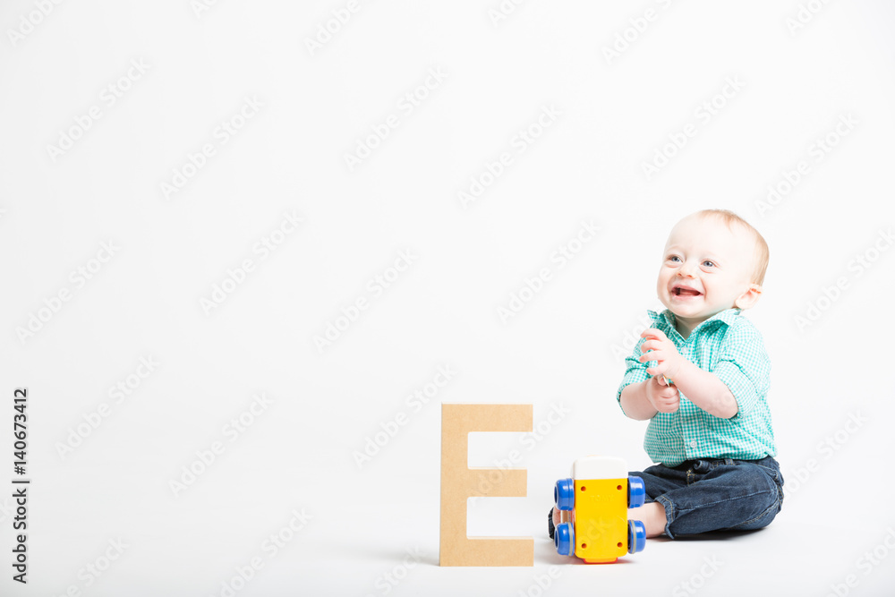 Baby Smiling on White with Wooden Letter and Toy. a 6 month old baby sitting in a white studio looking off camera next to a toy and a wooden letter e Baby Smiling on White with Wooden Letter and Toy. a 6 month old baby sitting in a white studio looking off camera next to a toy and a wooden letter e
