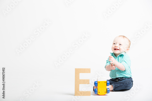 Baby Smiling on White with Wooden Letter and Toy. a 6 month old baby sitting in a white studio looking off camera next to a toy and a wooden letter e