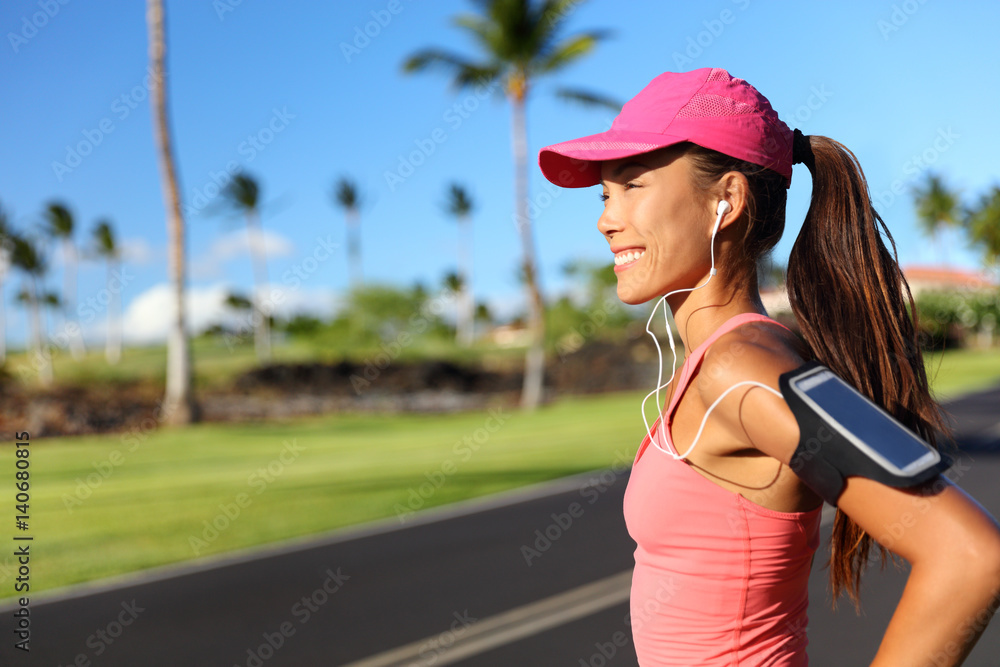Happy fitness girl runner listening to running motivation music on smartphone app with earphones. Asian woman athlete wearing cap and armband phone holder outdoor on city road.