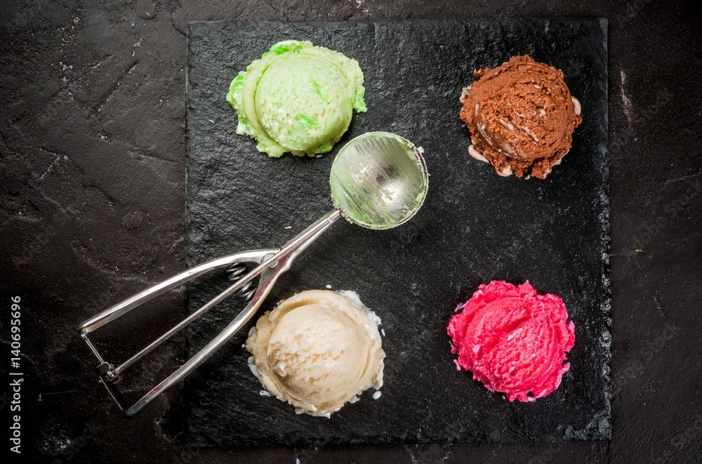 Selection of colorful home made ice cream: lemon (pistachio) green, berry pink, chocolate, white vanilla. With a spoon for serving balls, on a slate board, on a black table. Top view, copy space
