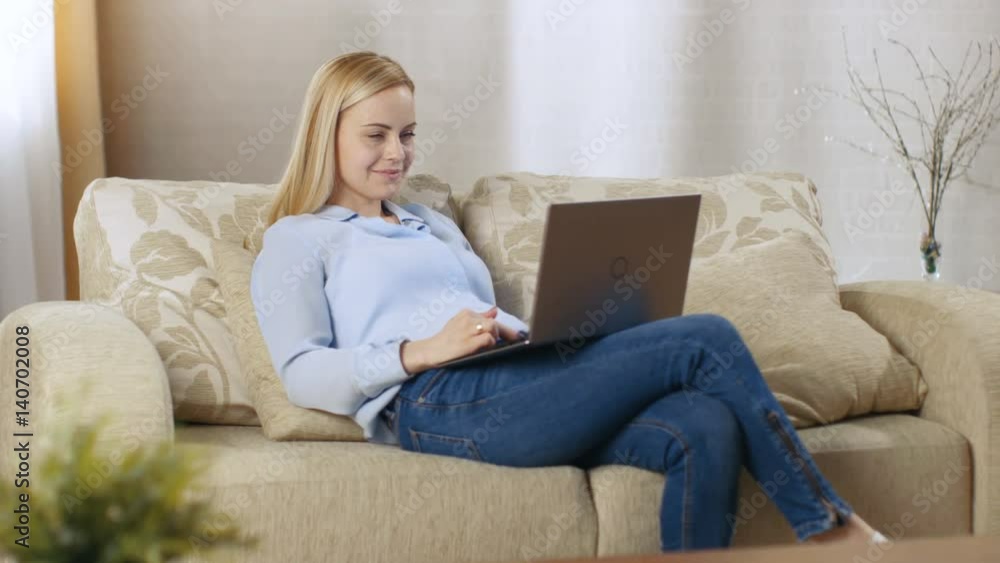 Beautiful Blonde Woman Sitting on a Couch with Notebook on Her Lap, She Actively Types on It. Her Room is Full of Light. Shot on RED EPIC-W 8K Helium Cinema Camera. Beautiful Blonde Woman Sitting on a Couch with Notebook on Her Lap, She Actively Types on It. Her Room is Full of Light. Shot on RED EPIC-W 8K Helium Cinema Camera.