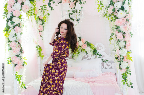 Charming young woman in a purple floral dress. She stands by the bed, decorated with flowers