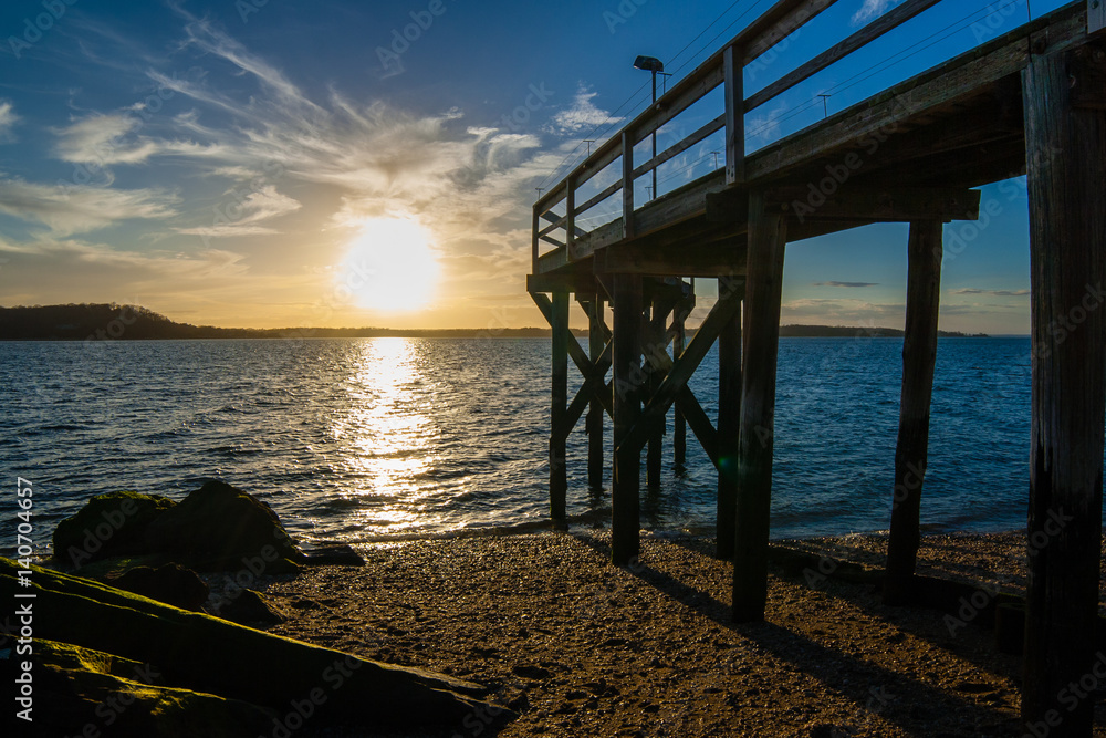 Sunset on a beach from between algae-covered rocks and a wooden pier. West Neck Beach in Huntington, NY, USA. Sunset on a beach from between algae-covered rocks and a wooden pier. West Neck Beach in Huntington, NY, USA.