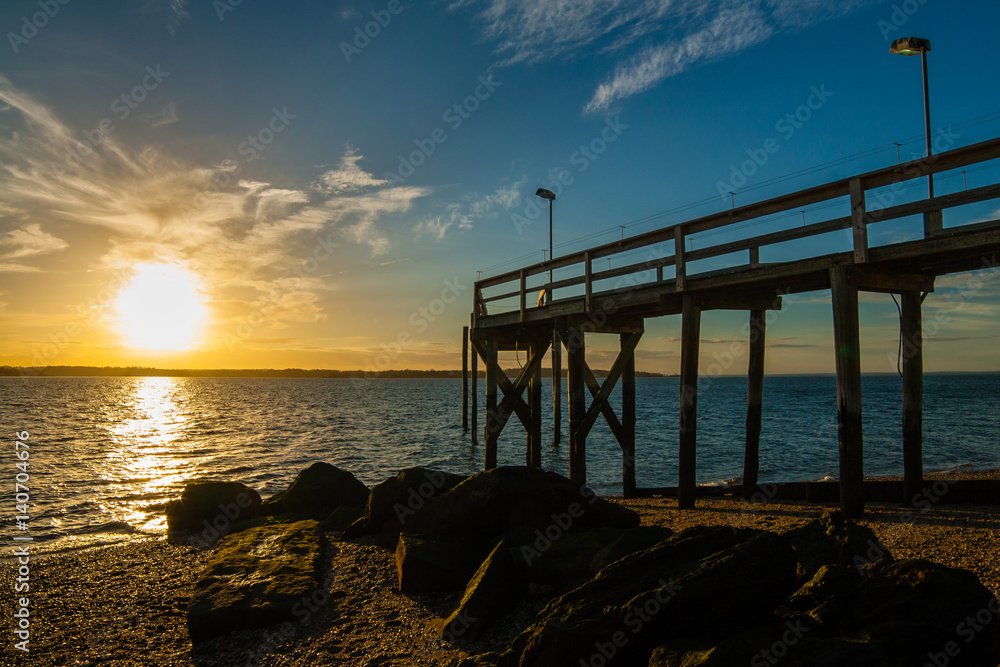 The setting sun with clouds in the background with a rocky beach, large algae-covered stones, and a wooden pier in the foreground. West Neck Beach in Huntington, NY, USA. The setting sun with clouds in the background with a rocky beach, large algae-covered stones, and a wooden pier in the foreground. West Neck Beach in Huntington, NY, USA.