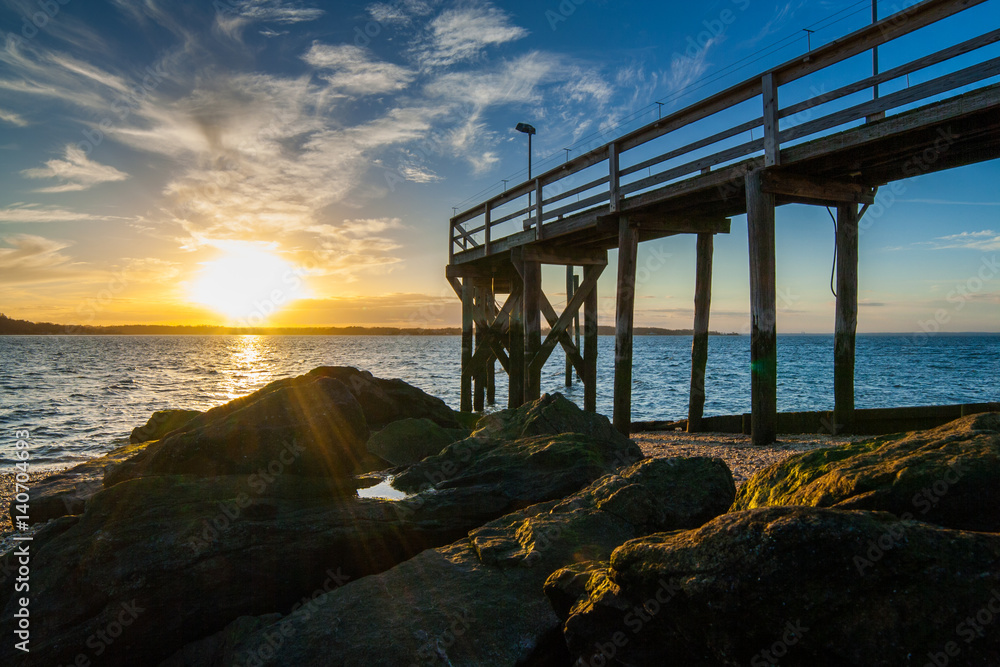 Naklejka premium The setting sun with clouds in the background with a rocky beach, large algae-covered stones, and a wooden pier in the foreground. West Neck Beach in Huntington, NY, USA.