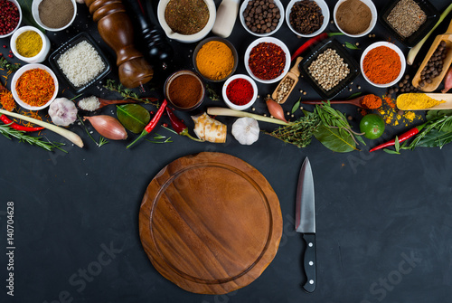 Large set of spices, herbs, vegetables, empty round cutting board and professional chef's knife on black background