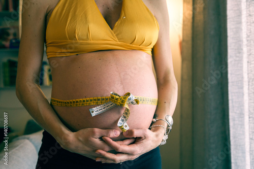 Belly of pregnant young woman with gift bow with tape measure. Prenatal health care concept