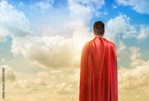 A businessman in super hero cape standing turned back on the sky background
