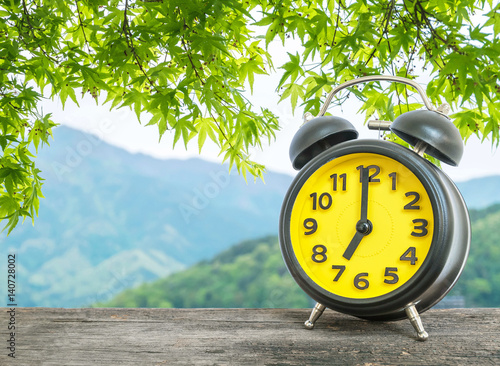 Closeup black and yellow alarm clock for decorate in 7 o'clock on blurred leaves and mountain view background