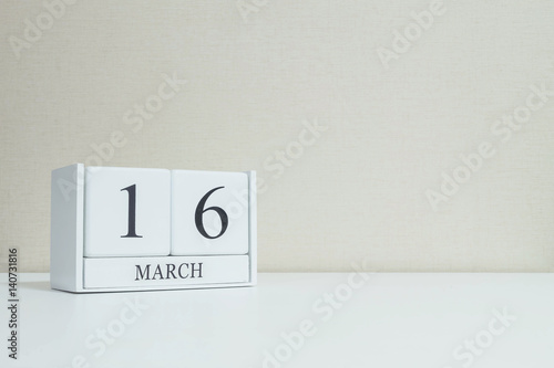 Closeup white wooden calendar with black 16 march word on blurred white wood desk and cream color wallpaper in room textured background with copy space , selective focus at the calendar