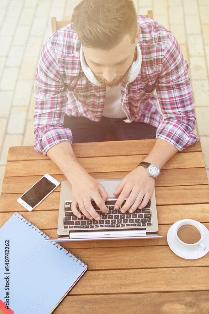 Businessman working outdoors on laptop and drinking a cup of coffee. Man with earphones sitting at the table in the cafe. Toned image. Businessman working outdoors on laptop and drinking a cup of coffee. Man with earphones sitting at the table in the cafe. Toned image.