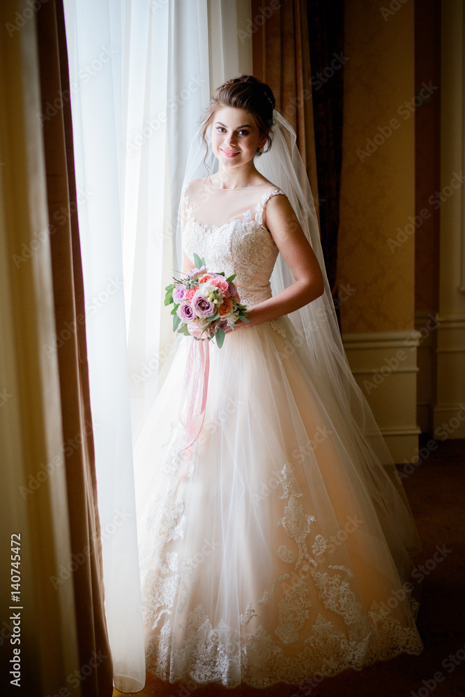 Bride with tender wedding bouquet of pink and violet flowers stands before the window